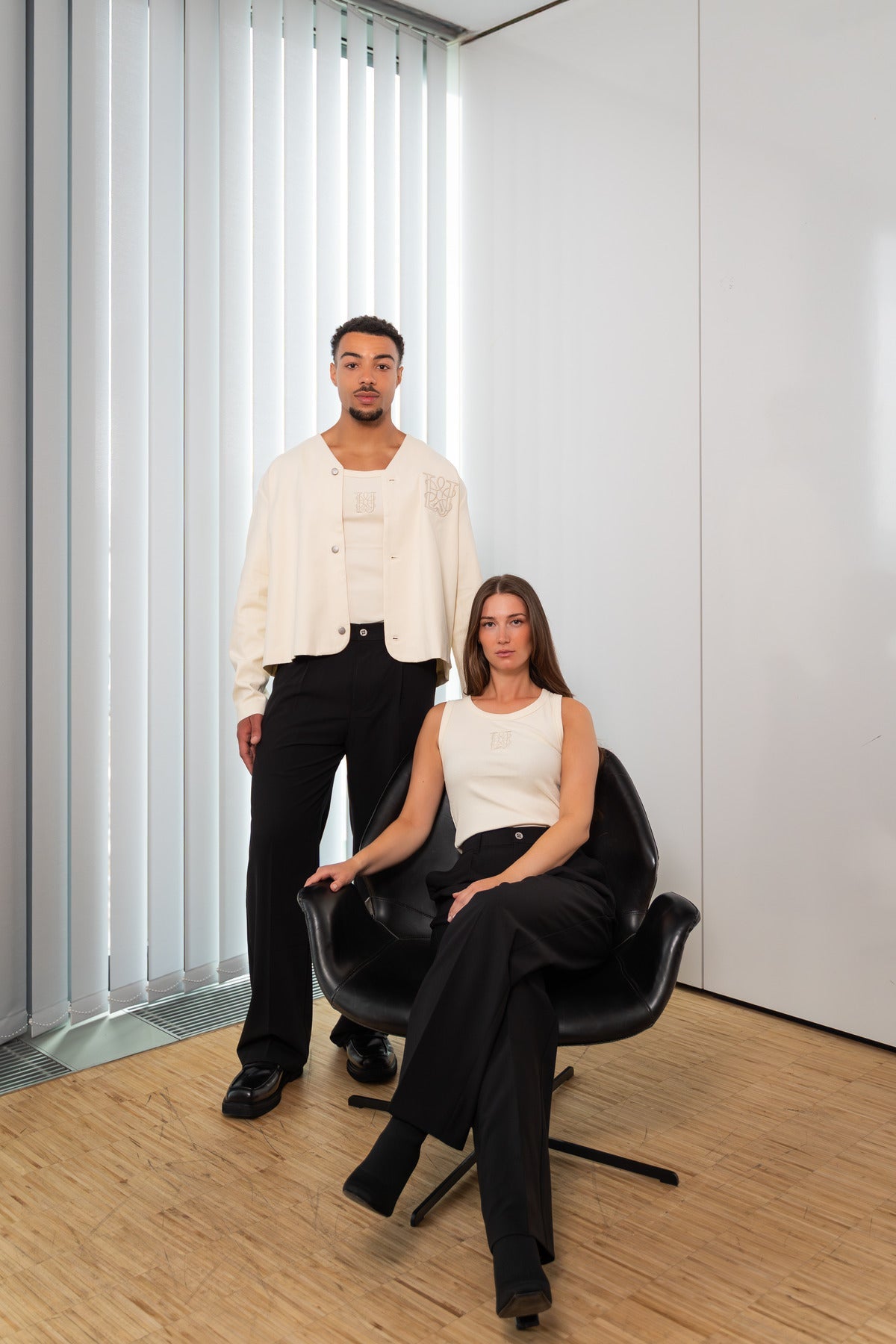Zwei Models in creme-schwarzen Outfits mit Monogramm-Details in einem modernen, hellen Raum mit Holzfußboden und weißen Jalousien.